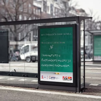 Bus stop shelter with a green chalkboard-style advertisement featuring Cyrillic text and logos at the bottom.