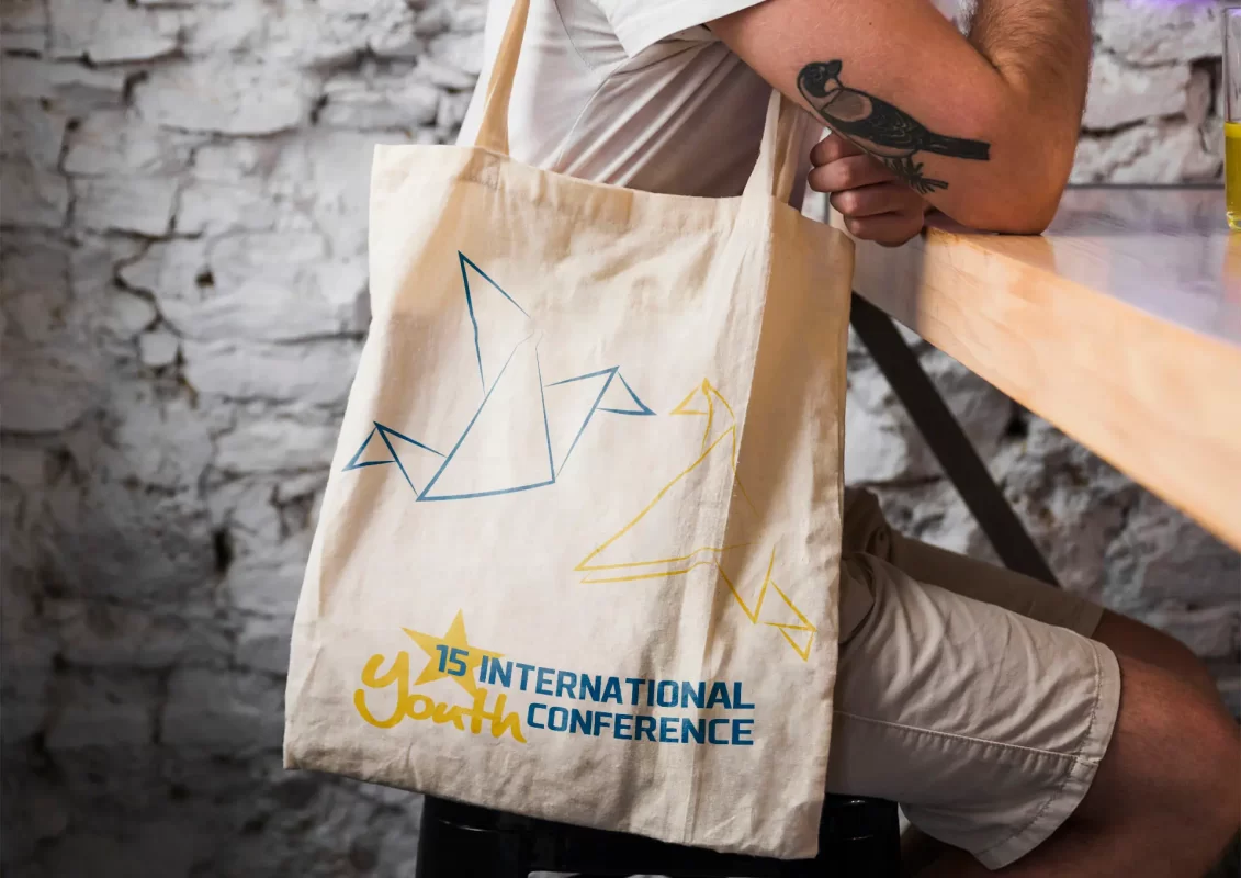 Person holding a tote bag with "15 International Youth Conference" and origami birds design.