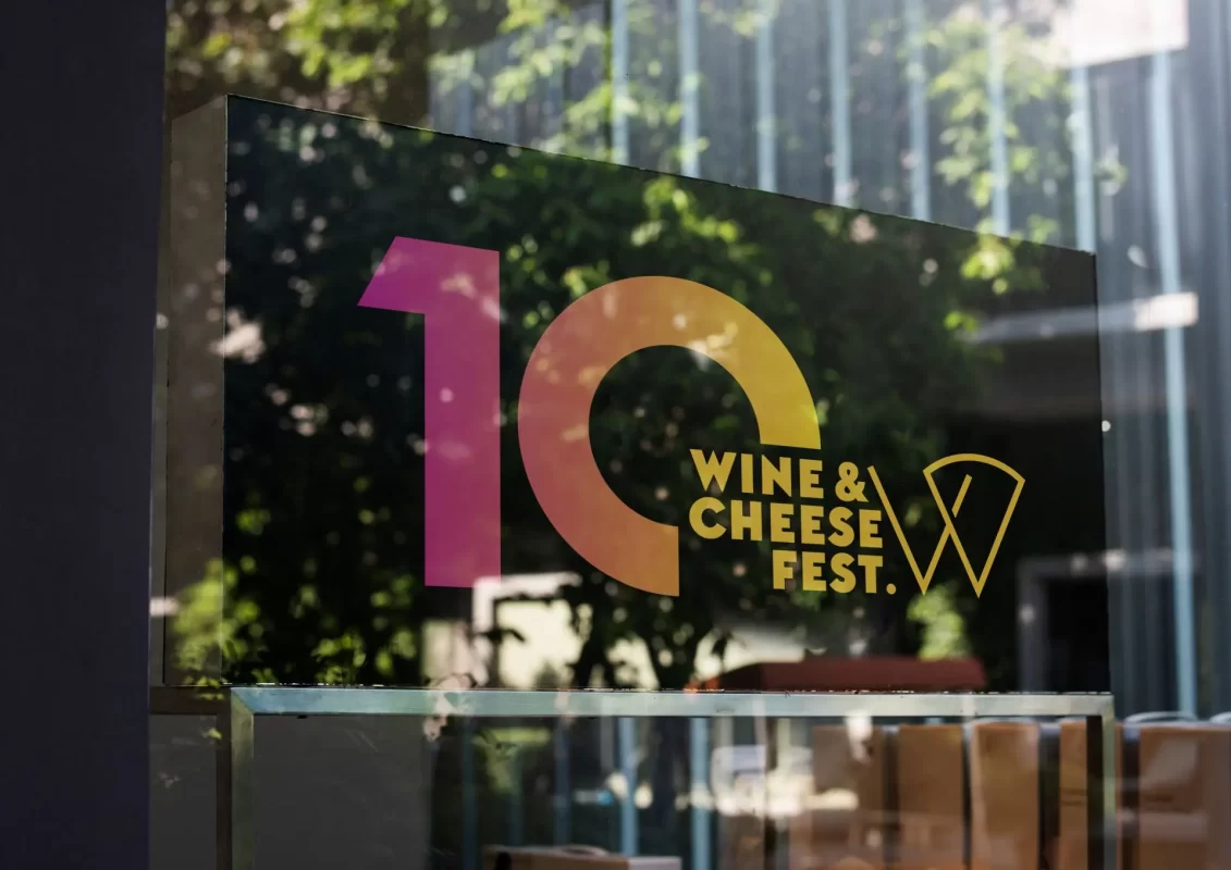 Glass sign with "10 Wine & Cheese Fest." logo in pink and gold, with blurred greenery background.