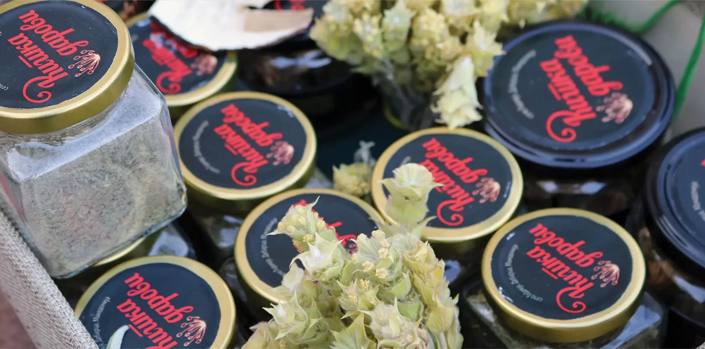 Jars of herbal tea with black lids, inscribed with red text, among dried flowers on a market stall.