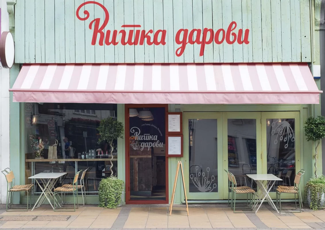 Shopfront of a café with pastel green walls, a pink-and-white striped awning, and the sign 'Китка дарови' in Cyrillic. There are outdoor tables, chairs, and potted plants near the entrance.