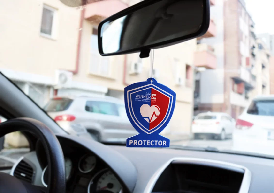 An air freshener hanging from a rearview mirror inside a car, with blurred street scene in background.