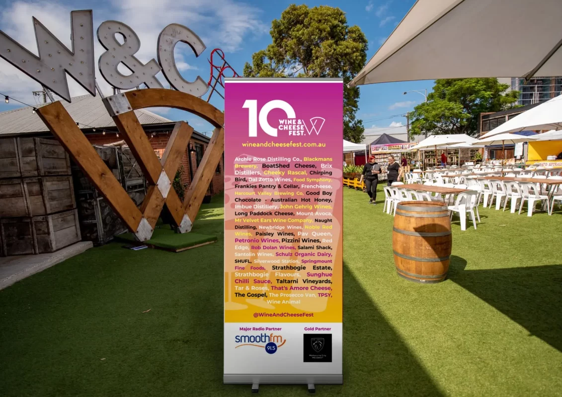 Outdoor festival with seating, a large "W&C" sculpture, and information banner for a wine and cheese fest.