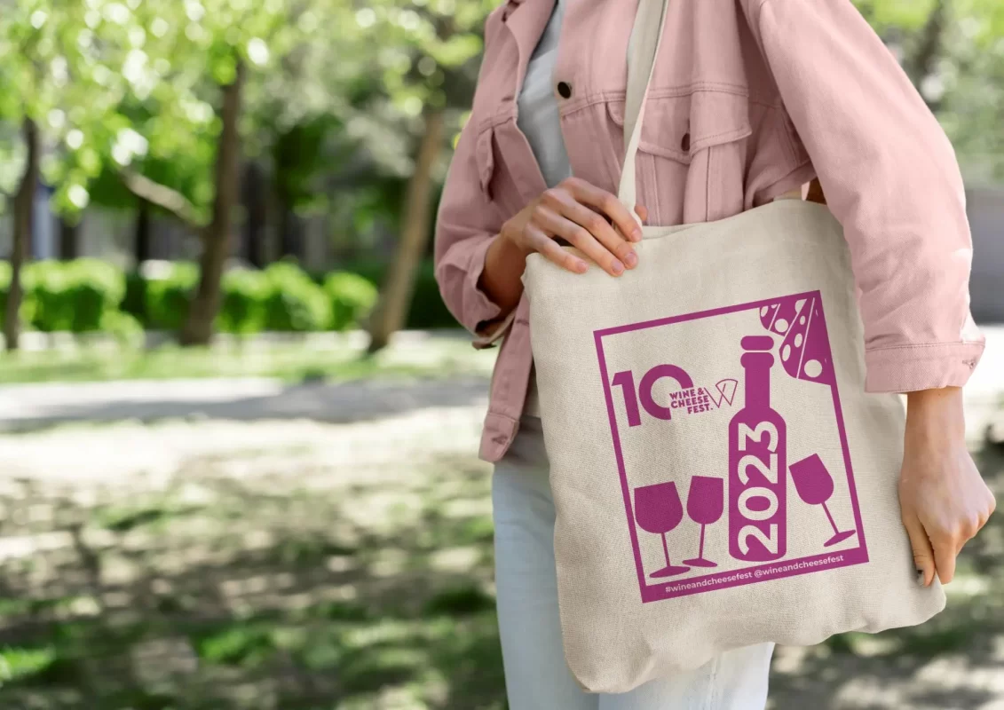 A person holding a tote bag with a 'Wine & Cheese Fest 2023' graphic in a park.